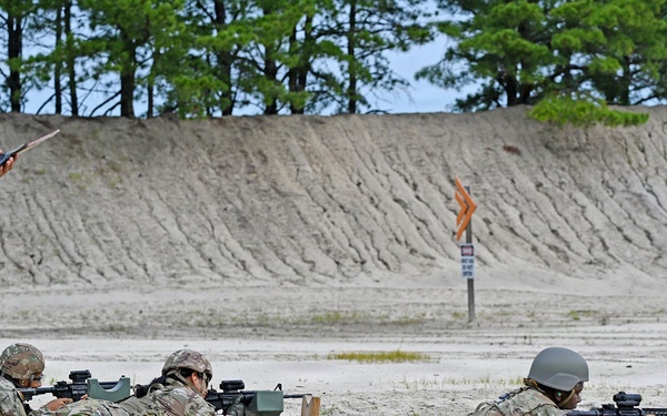 Military personnel are conducting a rifle qualification or marksmanship training exercise on a sandy firing range. Several soldiers are in the prone position aiming rifles downrange while a range safety officer or instructor wearing a high-visibility orange vest observes and records data on a tablet or clipboard. The setting appears to be a standard military live-fire range with a sand berm backstop.