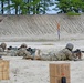 Military personnel are conducting a rifle qualification or marksmanship training exercise on a sandy firing range. Several soldiers are in the prone position aiming rifles downrange while a range safety officer or instructor wearing a high-visibility orange vest observes and records data on a tablet or clipboard. The setting appears to be a standard military live-fire range with a sand berm backstop.