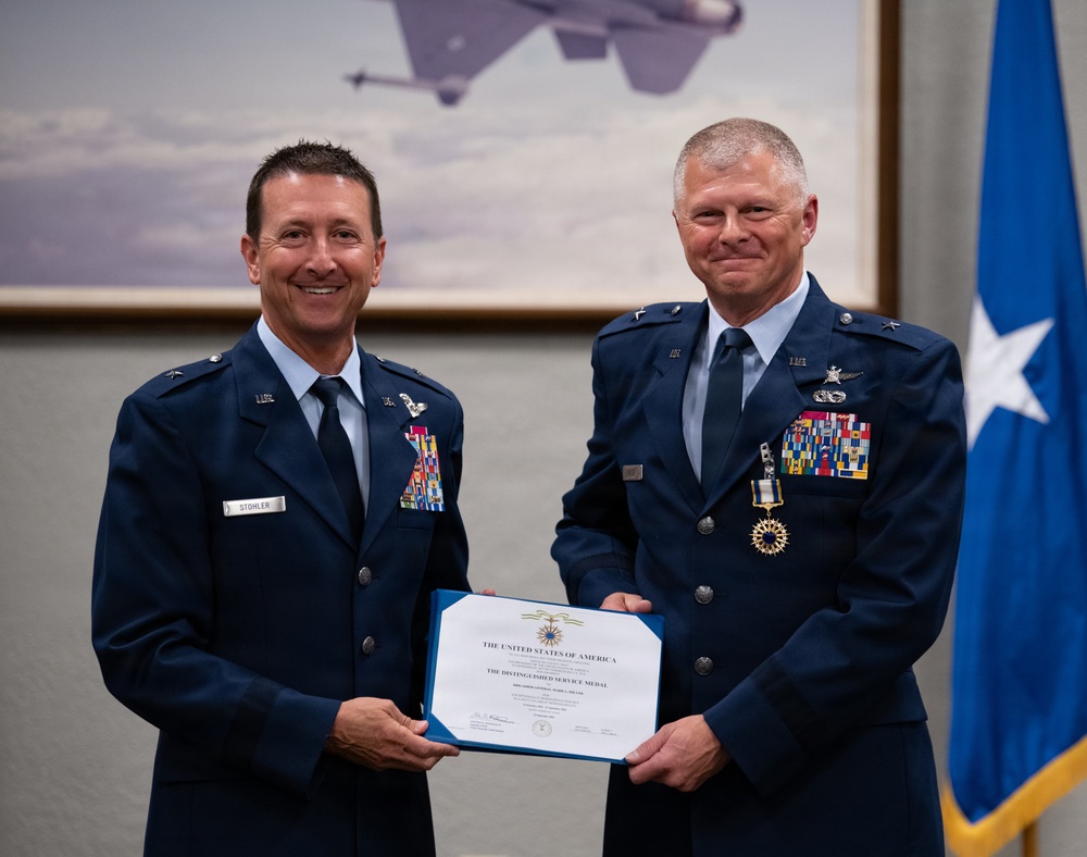 Two U.S. Air Force officers in dress blue uniforms pose together holding a Distinguished Service Medal certificate from the United States of America. The officer on the left, identified by his name tag as 'STOHLER,' appears to be presenting the award to the officer on the right, who is wearing the Distinguished Service Medal around his neck. The ceremony takes place in front of a military aviation painting and a one-star general officer flag.