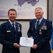Two U.S. Air Force officers in dress blue uniforms pose together holding a Distinguished Service Medal certificate from the United States of America. The officer on the left, identified by his name tag as 'STOHLER,' appears to be presenting the award to the officer on the right, who is wearing the Distinguished Service Medal around his neck. The ceremony takes place in front of a military aviation painting and a one-star general officer flag.