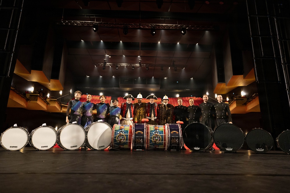 A group of approximately 12 performers pose on a theater stage with an array of bass drums in front of them. The group appears to consist of two distinct ensembles: one wearing colorful marching band-style uniforms with white plumed helmets, and another in all-black attire. The drums visible include Evans-branded white-headed bass drums and decorated traditional-style drums, suggesting a collaborative performance or competition setting.