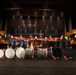 A group of approximately 12 performers pose on a theater stage with an array of bass drums in front of them. The group appears to consist of two distinct ensembles: one wearing colorful marching band-style uniforms with white plumed helmets, and another in all-black attire. The drums visible include Evans-branded white-headed bass drums and decorated traditional-style drums, suggesting a collaborative performance or competition setting.