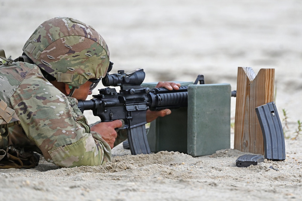 A soldier in MultiCam/OCP camouflage uniform is in a prone firing position on a sandy surface, aiming an M4/M16-style carbine equipped with a red dot or ACOG-type optic. The soldier is using a green military ammunition can as a rest/support for the weapon. Spare rifle magazines are visible nearby on a wooden magazine holder, indicating an active marksmanship training exercise.