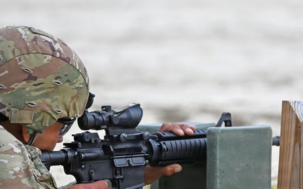A soldier in MultiCam/OCP camouflage uniform is in a prone firing position on a sandy surface, aiming an M4/M16-style carbine equipped with a red dot or ACOG-type optic. The soldier is using a green military ammunition can as a rest/support for the weapon. Spare rifle magazines are visible nearby on a wooden magazine holder, indicating an active marksmanship training exercise.