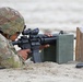 A soldier in MultiCam/OCP camouflage uniform is in a prone firing position on a sandy surface, aiming an M4/M16-style carbine equipped with a red dot or ACOG-type optic. The soldier is using a green military ammunition can as a rest/support for the weapon. Spare rifle magazines are visible nearby on a wooden magazine holder, indicating an active marksmanship training exercise.