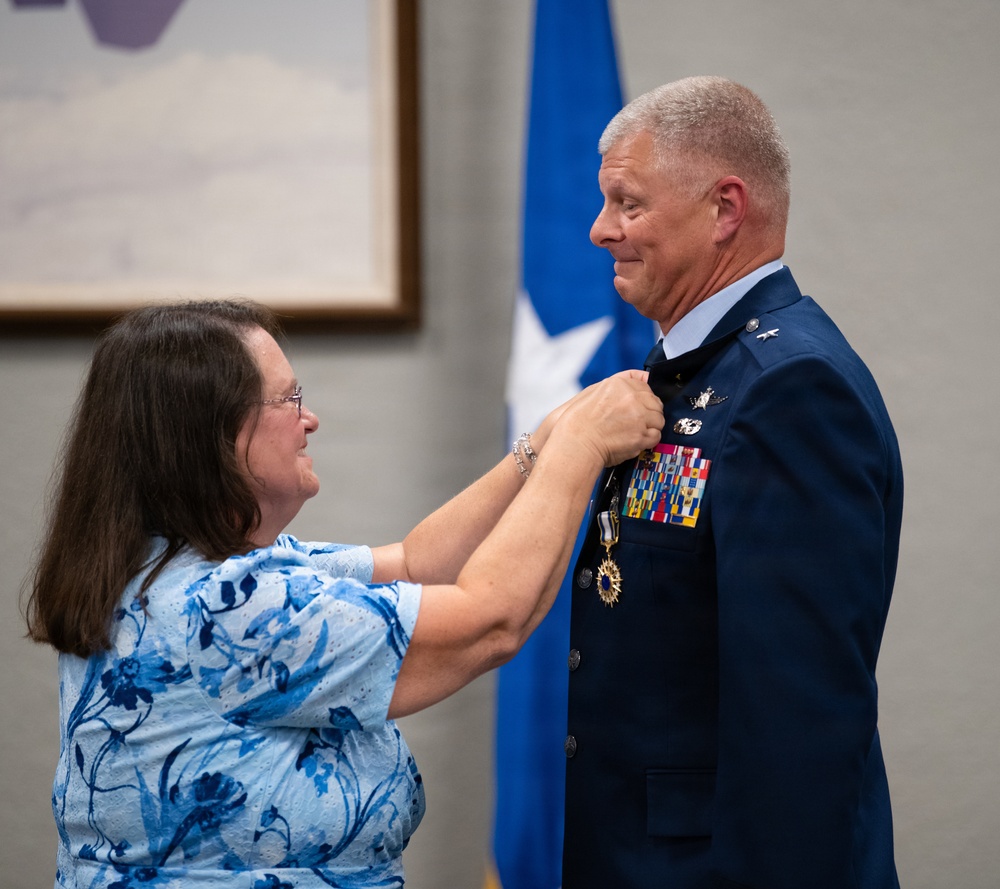 A woman in a blue floral top is pinning a medal or insignia onto the uniform of a male military officer in a dark blue Air Force dress uniform. The officer displays multiple service ribbons, medals, and rank insignia, with a one-star general flag visible in the background. This appears to be a formal military ceremony, likely a promotion, retirement, or decoration event.