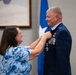 A woman in a blue floral top is pinning a medal or insignia onto the uniform of a male military officer in a dark blue Air Force dress uniform. The officer displays multiple service ribbons, medals, and rank insignia, with a one-star general flag visible in the background. This appears to be a formal military ceremony, likely a promotion, retirement, or decoration event.