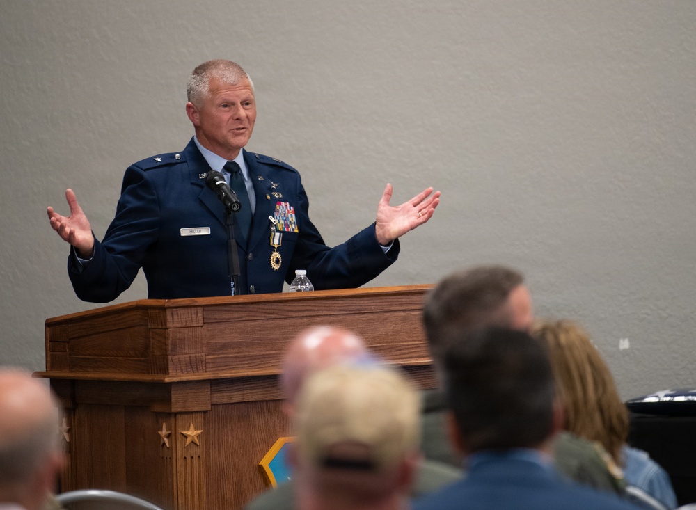 A U.S. Air Force officer, identified by his name tag as 'MILLER', is delivering a speech at a wooden podium to a seated audience. He is wearing a dress blue uniform with a general officer rank insignia (one star, indicating Brigadier General) and multiple service ribbons and medals. His hands are raised in an expressive gesture as he addresses the crowd.