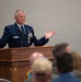 A U.S. Air Force officer, identified by his name tag as 'MILLER', is delivering a speech at a wooden podium to a seated audience. He is wearing a dress blue uniform with a general officer rank insignia (one star, indicating Brigadier General) and multiple service ribbons and medals. His hands are raised in an expressive gesture as he addresses the crowd.