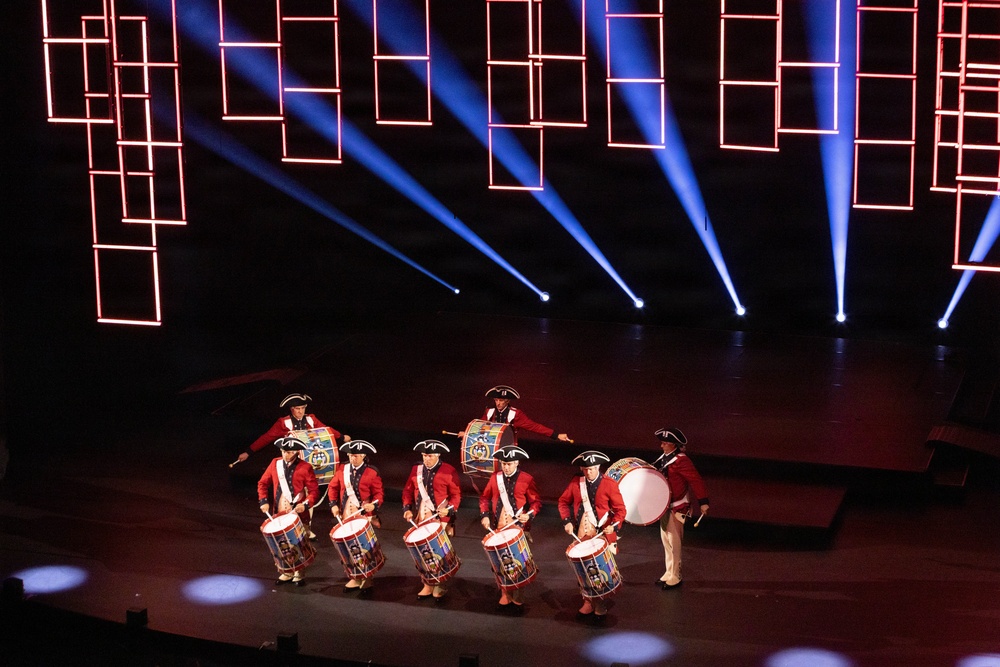 A drum corps performs on a darkened stage wearing colonial-era red uniforms and tricorn hats. The performance features approximately seven to eight drummers playing snare drums and a bass drum, set against a dramatic backdrop of blue spotlights and red rectangular neon light installations. The staging and costumes suggest a formal military tattoo or ceremonial performance event.