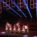 A drum corps performs on a darkened stage wearing colonial-era red uniforms and tricorn hats. The performance features approximately seven to eight drummers playing snare drums and a bass drum, set against a dramatic backdrop of blue spotlights and red rectangular neon light installations. The staging and costumes suggest a formal military tattoo or ceremonial performance event.