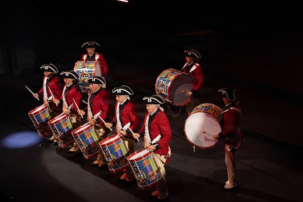 A group of performers dressed in 18th-century American Revolutionary War-era military uniforms are playing drums in a formal marching band or ceremonial performance. They wear distinctive red coats, tricorn hats with black and white cockades, and white breeches, consistent with Continental Army or Fife and Drum Corps attire. The performance appears to be taking place on a darkened stage or indoor venue with dramatic spotlight lighting.