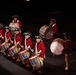 A group of performers dressed in 18th-century American Revolutionary War-era military uniforms are playing drums in a formal marching band or ceremonial performance. They wear distinctive red coats, tricorn hats with black and white cockades, and white breeches, consistent with Continental Army or Fife and Drum Corps attire. The performance appears to be taking place on a darkened stage or indoor venue with dramatic spotlight lighting.