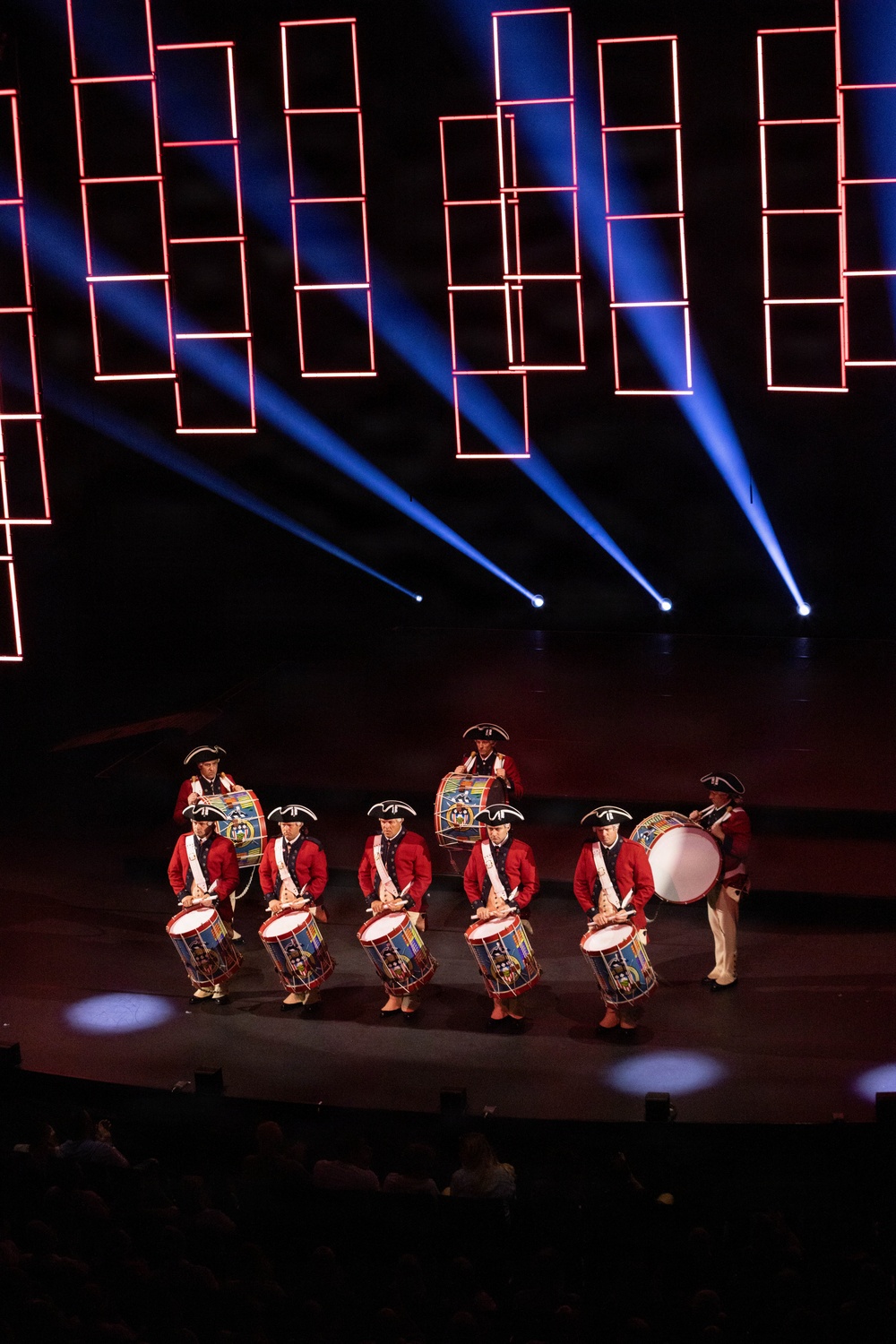 A drum corps performs on a darkened stage wearing 18th-century colonial-style military uniforms consisting of red coats, white cross-belts, and tricorn hats. The performers carry decorated snare drums and at least one large bass drum. The stage backdrop features illuminated red rectangular light frames and blue spotlight beams creating a dramatic visual effect.