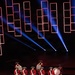A drum corps performs on a darkened stage wearing 18th-century colonial-style military uniforms consisting of red coats, white cross-belts, and tricorn hats. The performers carry decorated snare drums and at least one large bass drum. The stage backdrop features illuminated red rectangular light frames and blue spotlight beams creating a dramatic visual effect.