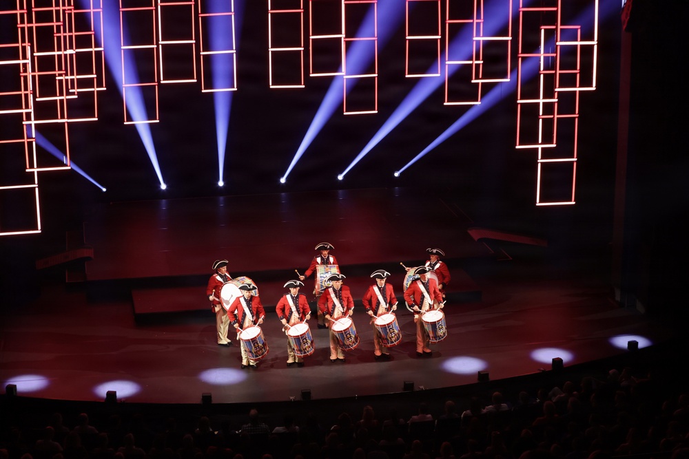 A group of performers dressed in colonial-era Revolutionary War uniforms — red coats, tricorn hats, and tan breeches — perform on a modern stage with dramatic lighting. They are playing snare drums and carrying a large bass drum, resembling a fife and drum corps. The stage features striking red neon rectangular light installations and blue spotlight beams against a dark backdrop.