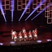 A group of performers dressed in colonial-era Revolutionary War uniforms — red coats, tricorn hats, and tan breeches — perform on a modern stage with dramatic lighting. They are playing snare drums and carrying a large bass drum, resembling a fife and drum corps. The stage features striking red neon rectangular light installations and blue spotlight beams against a dark backdrop.
