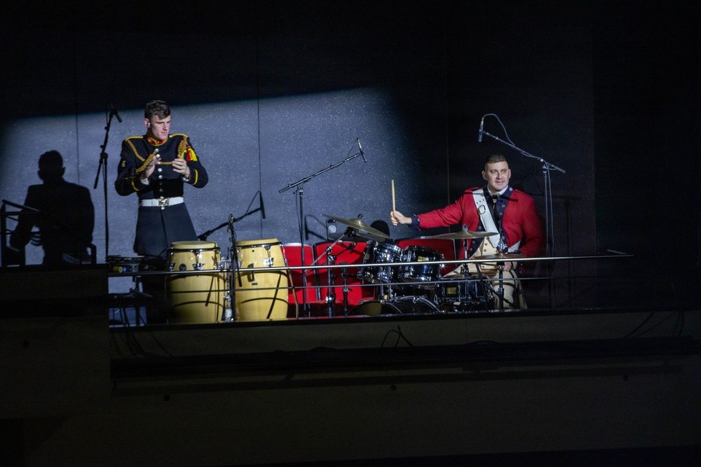 Two musicians in military dress uniforms perform on an elevated stage or balcony under dramatic spotlight lighting against a dark background. One musician in a dark uniform with gold trim plays a small hand percussion instrument, while another in a red jacket plays a full drum kit. The scene appears to be a formal military band or ceremonial performance.