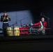 Two musicians in military dress uniforms perform on an elevated stage or balcony under dramatic spotlight lighting against a dark background. One musician in a dark uniform with gold trim plays a small hand percussion instrument, while another in a red jacket plays a full drum kit. The scene appears to be a formal military band or ceremonial performance.