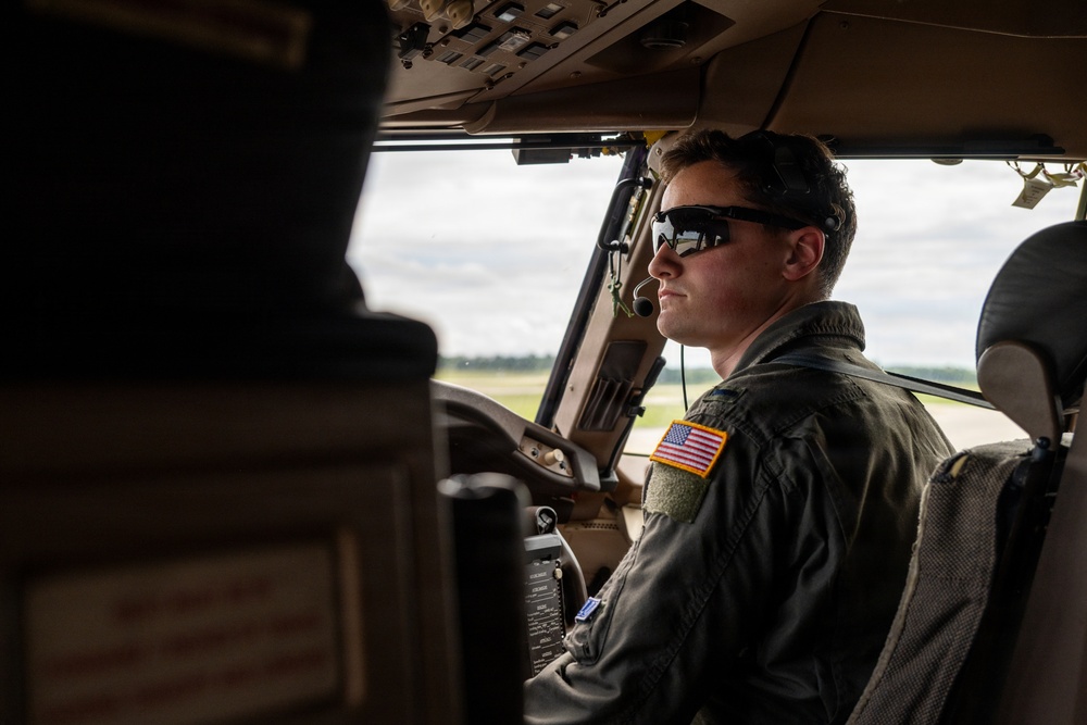 A U.S. military aviator is seated in the cockpit of a large aircraft, likely a transport or cargo plane, on or near a runway. The pilot is wearing a flight suit with a U.S. flag patch, sunglasses, and a communications headset. The image is taken from inside the cockpit looking toward the pilot from a rear-left angle.