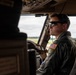 A U.S. military aviator is seated in the cockpit of a large aircraft, likely a transport or cargo plane, on or near a runway. The pilot is wearing a flight suit with a U.S. flag patch, sunglasses, and a communications headset. The image is taken from inside the cockpit looking toward the pilot from a rear-left angle.