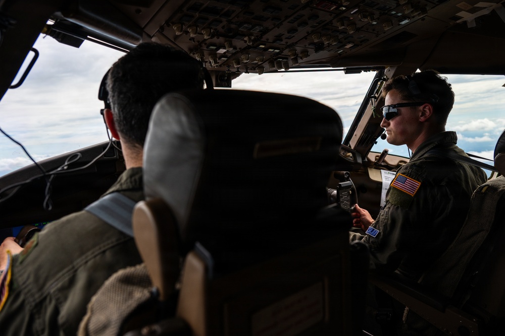 Two military aviators are seated in the cockpit of a large military aircraft in flight, with clouds visible through the windscreen. The pilot on the right is wearing sunglasses, a flight suit with a US flag patch, and a headset with boom microphone, and appears to be holding a checklist or reference card. The cockpit features an extensive overhead panel with numerous switches and controls, consistent with a large transport or tanker aircraft.