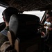 Two military aviators are seated in the cockpit of a large military aircraft in flight, with clouds visible through the windscreen. The pilot on the right is wearing sunglasses, a flight suit with a US flag patch, and a headset with boom microphone, and appears to be holding a checklist or reference card. The cockpit features an extensive overhead panel with numerous switches and controls, consistent with a large transport or tanker aircraft.