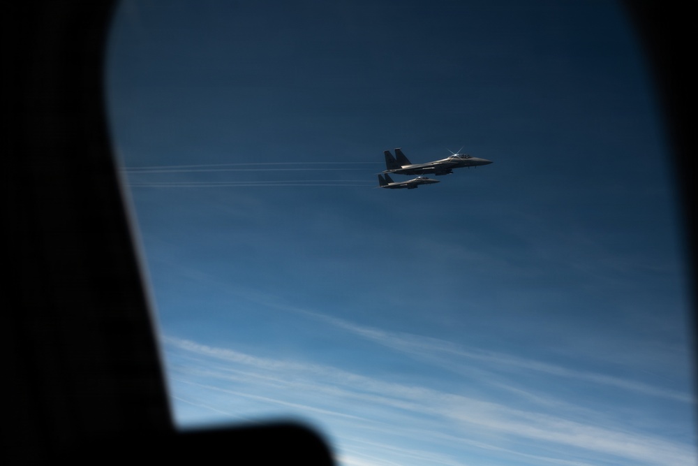 The image is taken from inside an aircraft through a window, showing two military jet fighters flying in close formation against a deep blue sky. The jets appear to be twin-tail fighter aircraft, consistent with F-15 Eagle variants, flying at altitude with contrail streaks visible in the background. The photo captures an escort or formation flight scenario from the perspective of a larger accompanying aircraft.