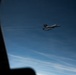 The image is taken from inside an aircraft through a window, showing two military jet fighters flying in close formation against a deep blue sky. The jets appear to be twin-tail fighter aircraft, consistent with F-15 Eagle variants, flying at altitude with contrail streaks visible in the background. The photo captures an escort or formation flight scenario from the perspective of a larger accompanying aircraft.