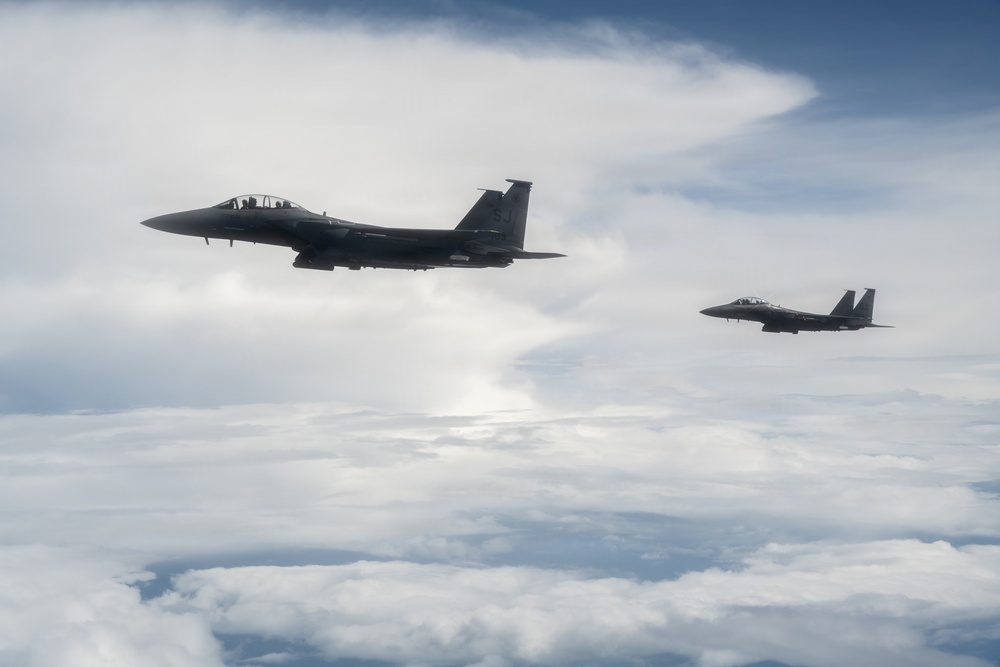 Two military fighter jets, identifiable as F-15 Eagles, are flying in formation above a cloud layer. The aircraft appear in a dark silhouette against an overcast sky with scattered cumulus clouds. The lead aircraft is prominently visible in the foreground while the second trails at a distance to the right.