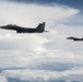 Two military fighter jets, identifiable as F-15 Eagles, are flying in formation above a cloud layer. The aircraft appear in a dark silhouette against an overcast sky with scattered cumulus clouds. The lead aircraft is prominently visible in the foreground while the second trails at a distance to the right.