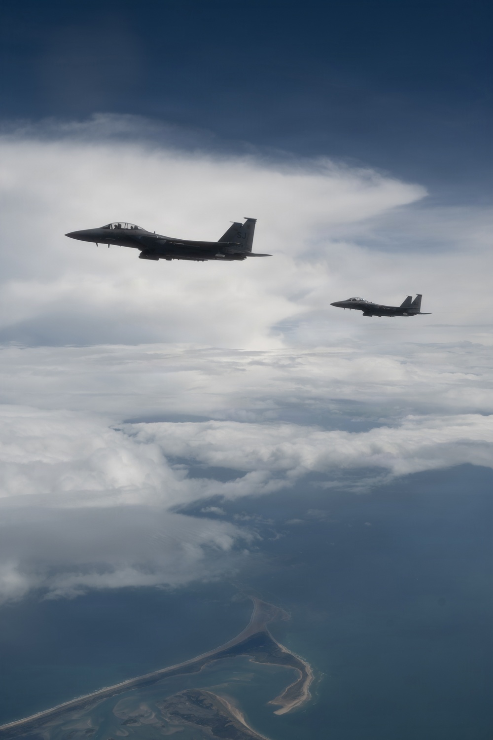Two military fighter jets, identifiable as F-15 Eagles, are flying in loose formation above a coastal landscape partially obscured by clouds. The aircraft are silhouetted against a partly cloudy sky, with a coastline and barrier island or peninsula visible far below. The image appears to be taken from a third aircraft flying alongside or slightly behind the pair.