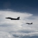 Two military fighter jets, identifiable as F-15 Eagles, are flying in loose formation above a coastal landscape partially obscured by clouds. The aircraft are silhouetted against a partly cloudy sky, with a coastline and barrier island or peninsula visible far below. The image appears to be taken from a third aircraft flying alongside or slightly behind the pair.