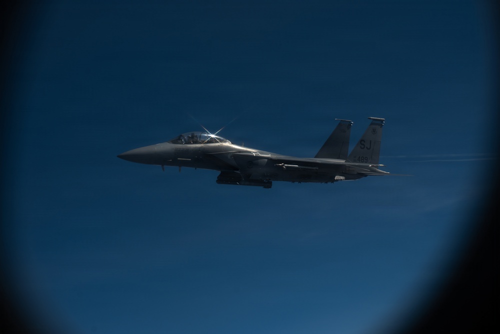 A U.S. Air Force F-15 Eagle fighter jet is photographed in flight against a deep blue sky. The aircraft bears the tail code 'SJ' and serial number '499', identifying it as belonging to Seymour Johnson Air Force Base in North Carolina. The image appears to have been taken from another aircraft, likely during a formation flight or aerial refueling operation.