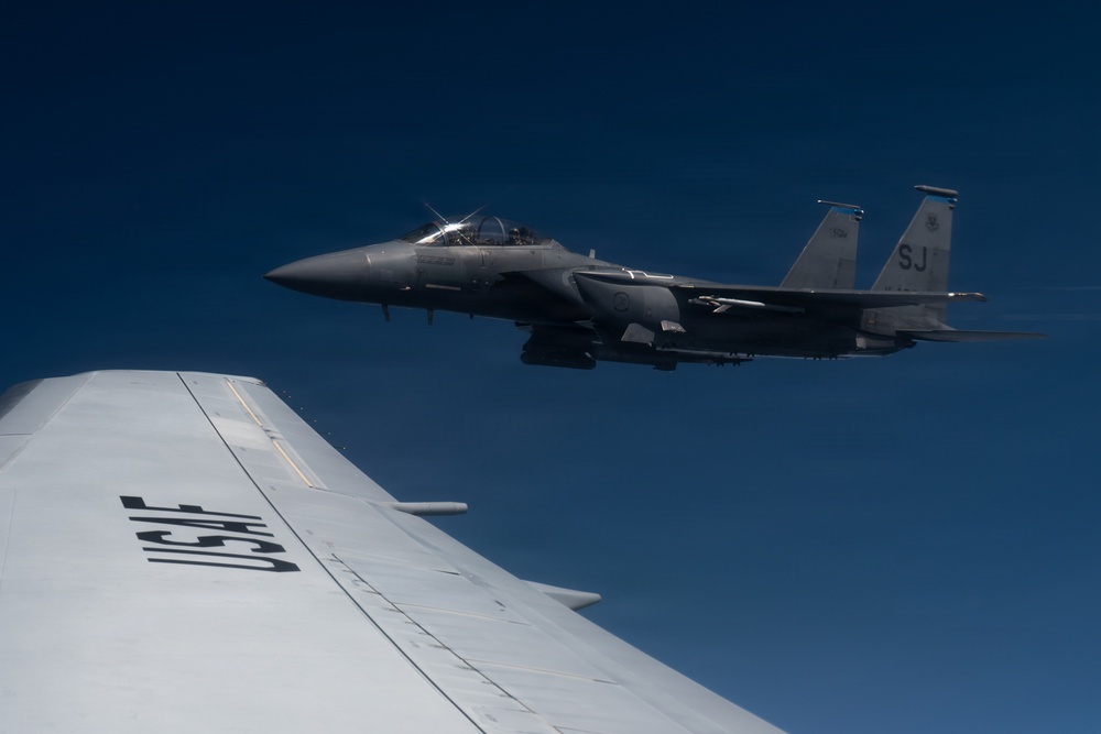 A U.S. Air Force F-15 Eagle fighter jet is flying in close formation alongside what appears to be a larger USAF aircraft, likely a tanker or transport, visible in the foreground. The F-15 bears the tail code 'SJ', indicating it belongs to Seymour Johnson Air Force Base in North Carolina. The image was taken from the wing or fuselage of the accompanying aircraft against a clear deep blue sky.