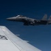 A U.S. Air Force F-15 Eagle fighter jet is flying in close formation alongside what appears to be a larger USAF aircraft, likely a tanker or transport, visible in the foreground. The F-15 bears the tail code 'SJ', indicating it belongs to Seymour Johnson Air Force Base in North Carolina. The image was taken from the wing or fuselage of the accompanying aircraft against a clear deep blue sky.
