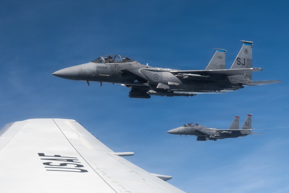 Two U.S. Air Force F-15 Strike Eagle fighter jets bearing the tail code 'SJ' (Seymour Johnson Air Force Base) fly in formation alongside a larger USAF aircraft, likely a tanker or transport. The image is taken from the wing or fuselage of the accompanying USAF aircraft, with 'USAF' markings clearly visible in the foreground. Both F-15s appear to be carrying external stores and are flying in clear blue sky conditions.