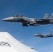 Two U.S. Air Force F-15 Strike Eagle fighter jets bearing the tail code 'SJ' (Seymour Johnson Air Force Base) fly in formation alongside a larger USAF aircraft, likely a tanker or transport. The image is taken from the wing or fuselage of the accompanying USAF aircraft, with 'USAF' markings clearly visible in the foreground. Both F-15s appear to be carrying external stores and are flying in clear blue sky conditions.