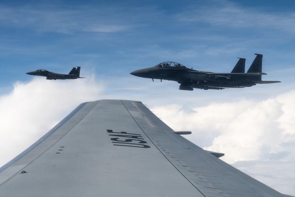 Two U.S. Air Force F-15 Eagle fighter jets are flying in formation alongside a larger USAF aircraft, photographed from the wing of what appears to be a transport or tanker aircraft. The 'USAF' marking is clearly visible on the foreground wing. The aircraft are flying above a cloud layer under a partly cloudy blue sky.