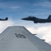Two U.S. Air Force F-15 Eagle fighter jets are flying in formation alongside a larger USAF aircraft, photographed from the wing of what appears to be a transport or tanker aircraft. The 'USAF' marking is clearly visible on the foreground wing. The aircraft are flying above a cloud layer under a partly cloudy blue sky.