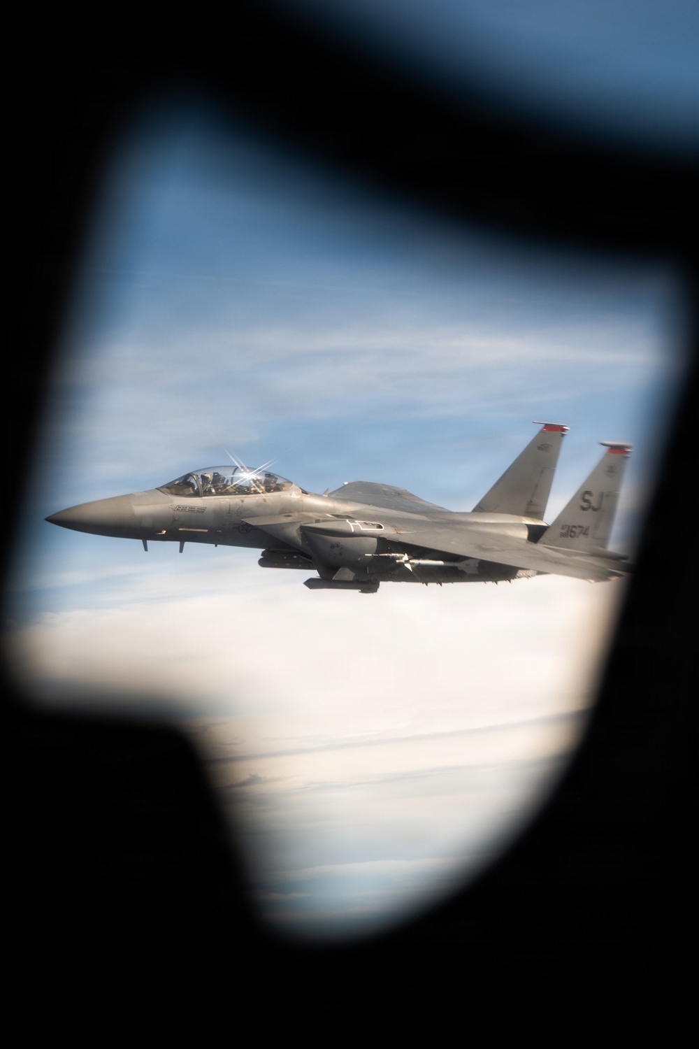 A U.S. Air Force F-15 Strike Eagle is photographed in flight through what appears to be a window or porthole of another aircraft. The jet bears tail code 'SJ' and serial number ending in '3674', indicating assignment to Seymour Johnson Air Force Base. The aircraft is flying at altitude with a blue sky and cloud layer visible in the background.