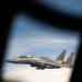 A U.S. Air Force F-15 Strike Eagle is photographed in flight through what appears to be a window or porthole of another aircraft. The jet bears tail code 'SJ' and serial number ending in '3674', indicating assignment to Seymour Johnson Air Force Base. The aircraft is flying at altitude with a blue sky and cloud layer visible in the background.