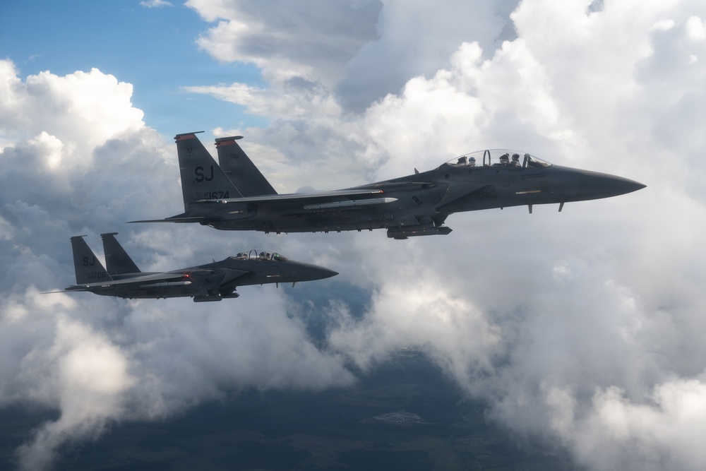 Two U.S. Air Force F-15E Strike Eagle fighter jets are flying in formation above a cloud layer. Both aircraft bear the 'SJ' tail code, indicating they belong to the 4th Fighter Wing based at Seymour Johnson Air Force Base, North Carolina. The foreground aircraft displays the serial number 11674 and appears to be carrying external ordnance or fuel tanks on its hardpoints.