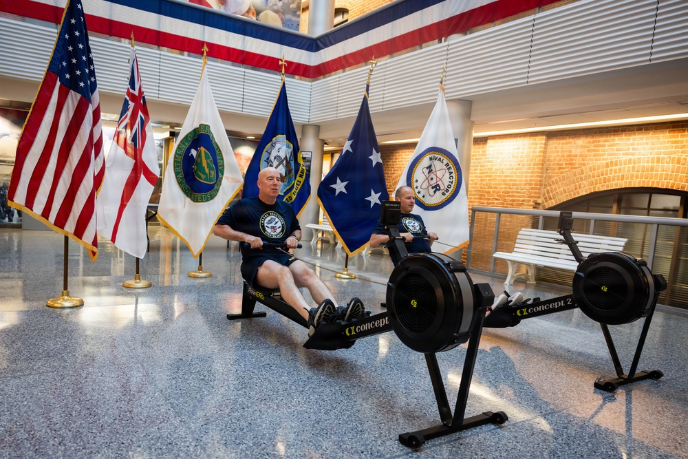 Two men in matching dark Navy-branded athletic wear are using Concept2 rowing machines in what appears to be a government or military building lobby. The setting is decorated with multiple flags including the U.S. flag, a British naval ensign, Department of Energy flag, U.S. Navy flag, a flag with a star (possibly flag officer pennant), and a Naval Reactors flag. The event appears to be an organized fitness demonstration or charity rowing event in an official military/government facility.