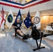 Two men in matching dark Navy-branded athletic wear are using Concept2 rowing machines in what appears to be a government or military building lobby. The setting is decorated with multiple flags including the U.S. flag, a British naval ensign, Department of Energy flag, U.S. Navy flag, a flag with a star (possibly flag officer pennant), and a Naval Reactors flag. The event appears to be an organized fitness demonstration or charity rowing event in an official military/government facility.