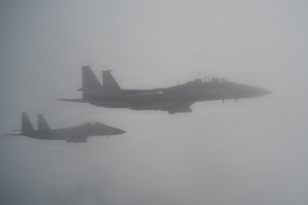 Two military fighter jets, consistent with F-15 Eagle aircraft, are flying in formation through heavy overcast or foggy conditions. The aircraft appear to be flying in a loose echelon or trail formation with the lead aircraft positioned higher and to the right. Visibility is significantly reduced due to atmospheric haze or cloud cover.