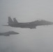 Two military fighter jets, consistent with F-15 Eagle aircraft, are flying in formation through heavy overcast or foggy conditions. The aircraft appear to be flying in a loose echelon or trail formation with the lead aircraft positioned higher and to the right. Visibility is significantly reduced due to atmospheric haze or cloud cover.