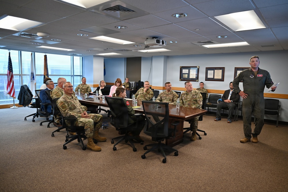 A military briefing or meeting is taking place in a formal conference room, with one individual in a flight suit standing and presenting to a group of seated military personnel and civilians. The majority of attendees are wearing OCP (Operational Camouflage Pattern) uniforms, suggesting a U.S. Army or Air Force context. The setting appears to be an official military facility based on the framed documents, flags, and institutional furnishings.