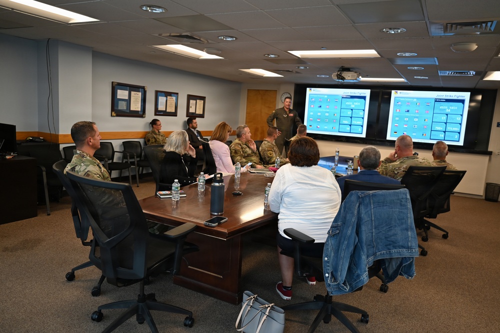 A military briefing is taking place in a conference room where a presenter in a flight suit stands before a large display screen showing slides titled 'Joint Strike Fighter.' A mixed group of military personnel in camouflage uniforms and civilians are seated around a large wooden conference table. The setting appears to be an official military facility based on the framed documents/awards on the wall and the formal briefing format.
