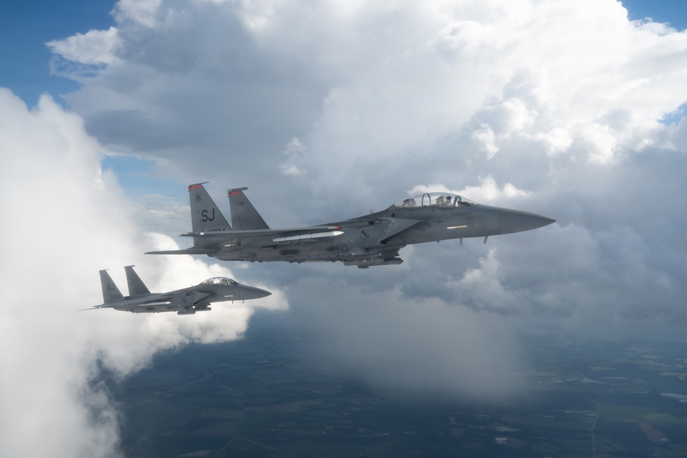 Two U.S. Air Force F-15E Strike Eagle fighter jets are flying in formation above cloud cover. The lead aircraft displays 'SJ' tail codes, identifying it as belonging to the 4th Fighter Wing based at Seymour Johnson Air Force Base, North Carolina. Both aircraft appear to be carrying external ordnance and fuel configurations on their hardpoints.